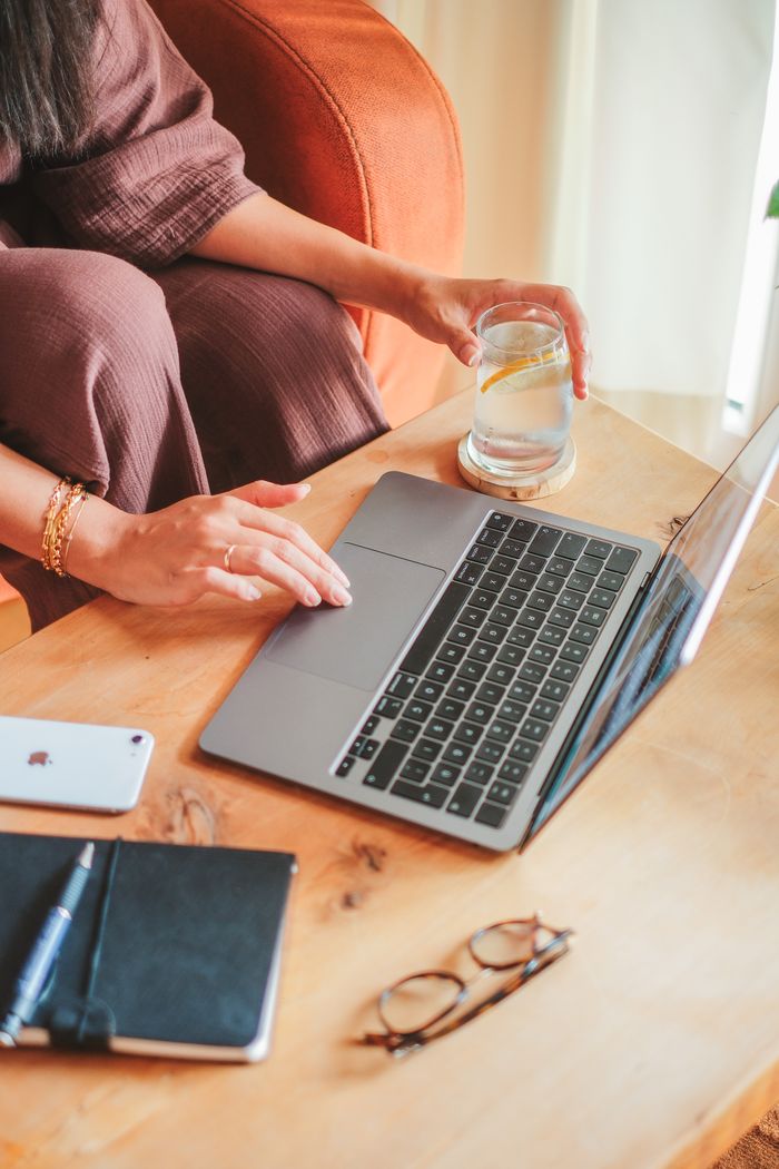 Woman using a laptop at home with a glass of lemon water, smartphone, and notebook on the table.